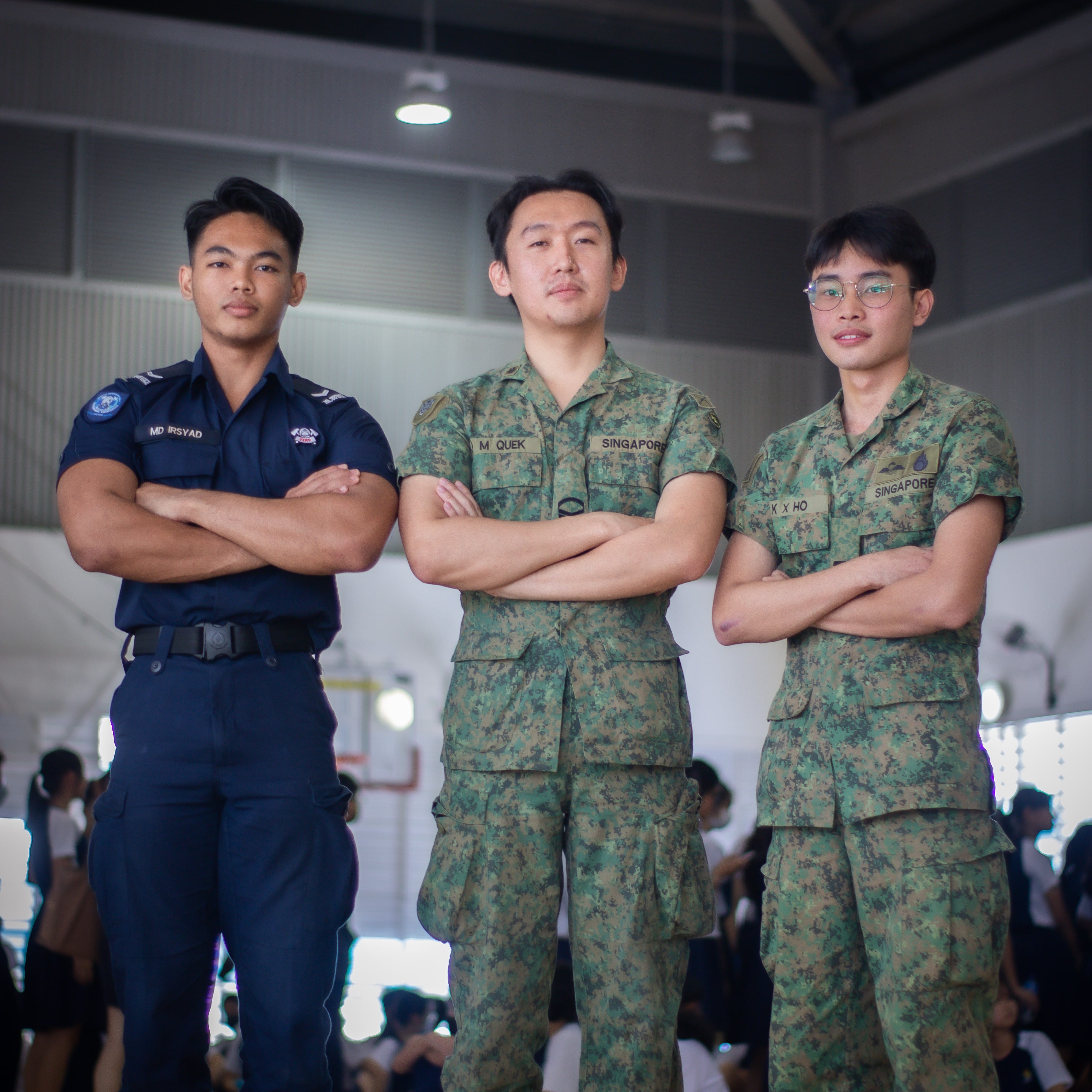 Three young men in Singapore military and police uniforms stand with arms crossed indoors.